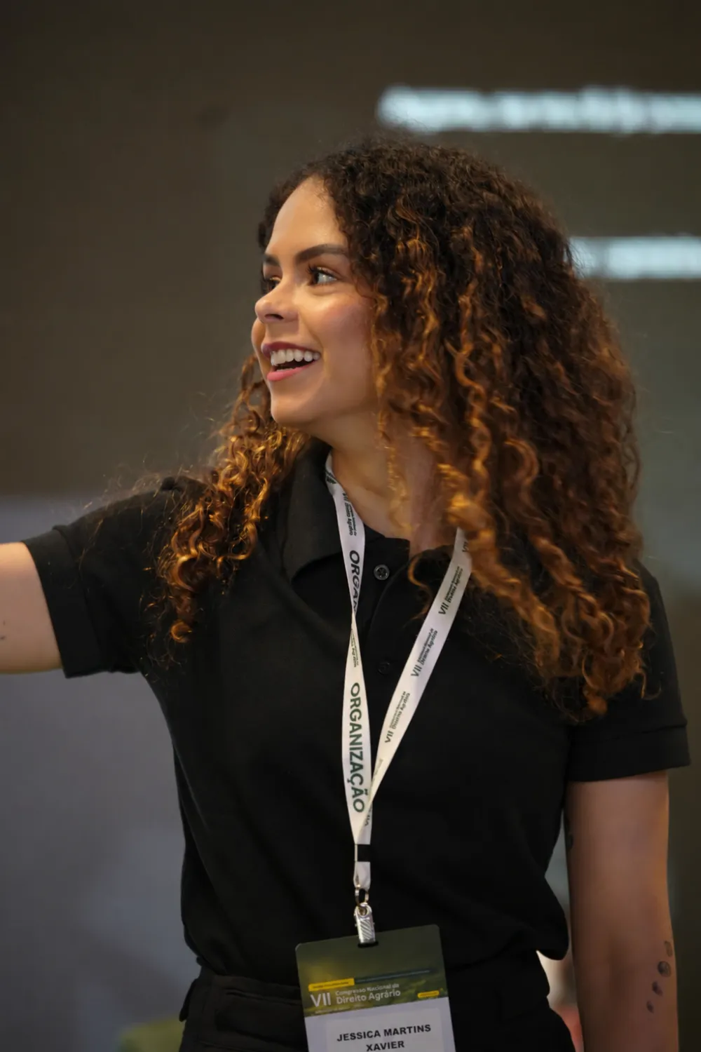 Jéssica Xavier sorrindo em um evento, vestindo camisa preta com credencial de organização